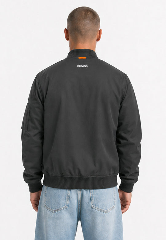 Eine Person mit kurzen Haaren, die die anthrazitfarbene Racing Jacke RECARO aus dem RECARO Shop trägt – eine wind- und wasserabweisende Jacke im Bomber-Stil – sowie hellblaue Jeans, steht mit dem Rücken zur Kamera vor einem schlichten weißen Hintergrund.