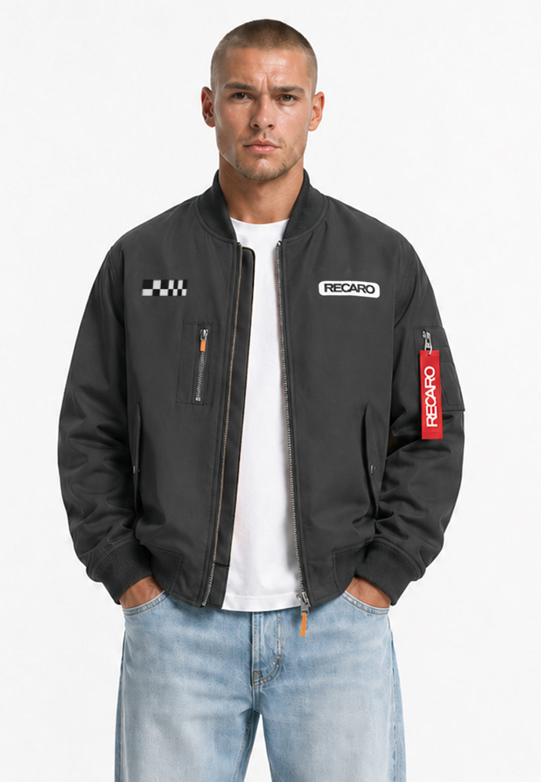 Ein Mann, der eine anthrazitfarbene RECARO Shop Racing Jacke mit klassischer Bomber-Silhouette und markanten RECARO-Aufnähern trägt, steht mit den Händen in den Taschen vor einem schlichten weißen Hintergrund. Kombiniert mit einem weißen T-Shirt und hellblauen Jeans bietet das wind- und wasserabweisende Material der Jacke sowohl Stil als auch Funktionalität.