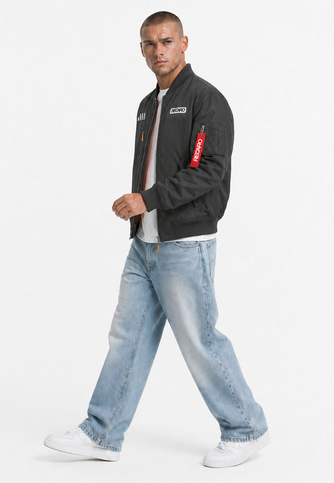 Ein Mann, der eine schwarze RECARO-Rennjacke in Anthrazit aus dem RECARO-Shop, ein weißes T-Shirt, hellblaue Baggy-Jeans und weiße Sneaker trägt, geht vor einem schlichten weißen Hintergrund voran. Das wind- und wasserabweisende Material seiner Jacke rundet den Look stilvoll und funktional ab.