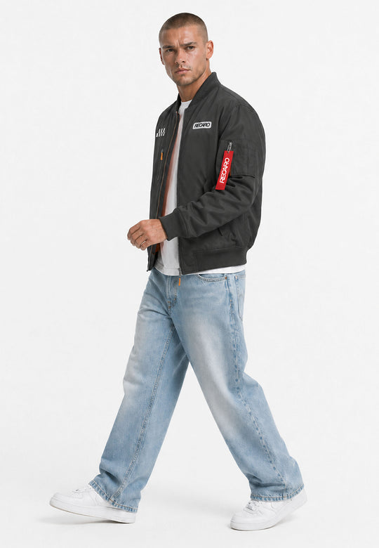 Ein Mann, der eine schwarze RECARO-Rennjacke in Anthrazit aus dem RECARO-Shop, ein weißes T-Shirt, hellblaue Baggy-Jeans und weiße Sneaker trägt, geht vor einem schlichten weißen Hintergrund voran. Das wind- und wasserabweisende Material seiner Jacke rundet den Look stilvoll und funktional ab.