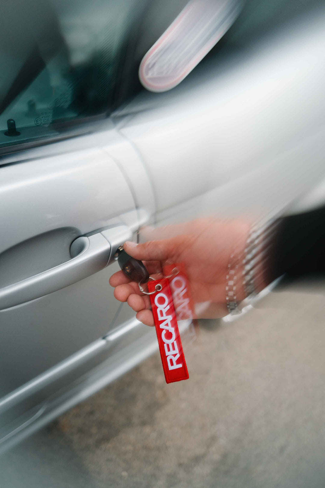 Eine Hand, an der die Jacke „Racing Jacke RECARO“ in Anthrazit aus dem RECARO Shop sitzt – eine Jacke im Bomber-Stil, die sowohl wind- als auch wasserabweisend ist –, öffnet mit einem Schlüssel, an dem ein roter RECARO-Schlüsselanhänger hängt, eine silberne Autotür, während sie eine Metalluhr trägt.