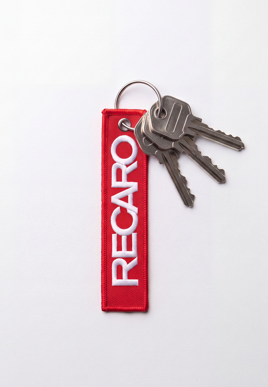 Der Schlüsselanhänger RECARO Textil von RECARO Shop besteht aus vier silbernen Schlüsseln an einem Schlüsselring mit einem roten Stoffband, auf dem in weißer Schrift RECARO auf weißem Hintergrund steht.