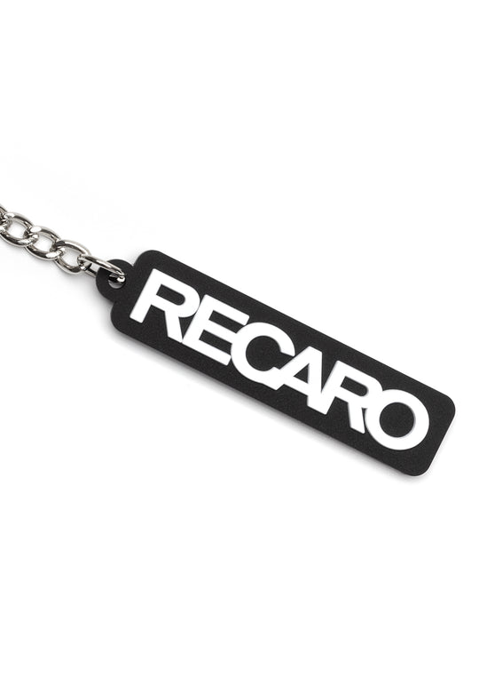 Der Schlüsselanhänger Originals RECARO aus dem RECARO Shop ist ein schwarzer, rechteckiger Gummischlüsselanhänger mit auffälligem, weißem Schriftzug, der an einer silbernen Metallkette befestigt ist und auf weißem Hintergrund für sportlichen Style sorgt.