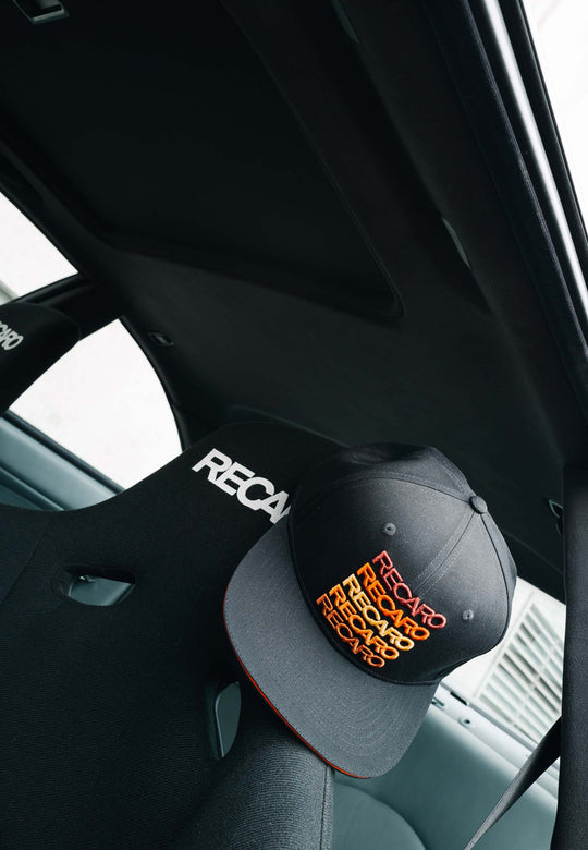 Eine schwarz-orangefarbene Snapback-Kappe „Spektrum“ von RECARO_Merchandising im Retro-Look liegt auf der Kopfstütze eines schwarzen RECARO-Autositzes, gesehen aus dem Fahrzeuginneren.