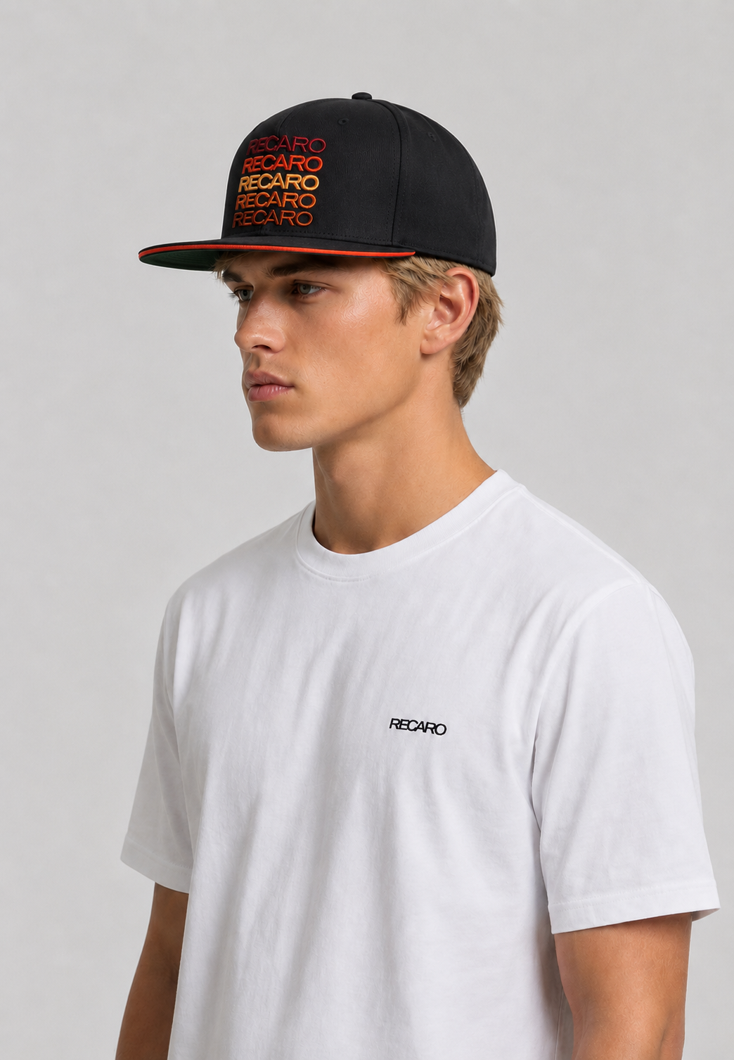 Ein junger Mann in einem weißen T-Shirt und der RECARO_Merchandising Snapback-Kappe „Spektrum“ in Schwarz-Orange mit 3D-Farbspektrum-Stickerei blickt vor einem schlichten grauen Hintergrund zur Seite und verströmt dabei einen unverwechselbaren Retro-Look.