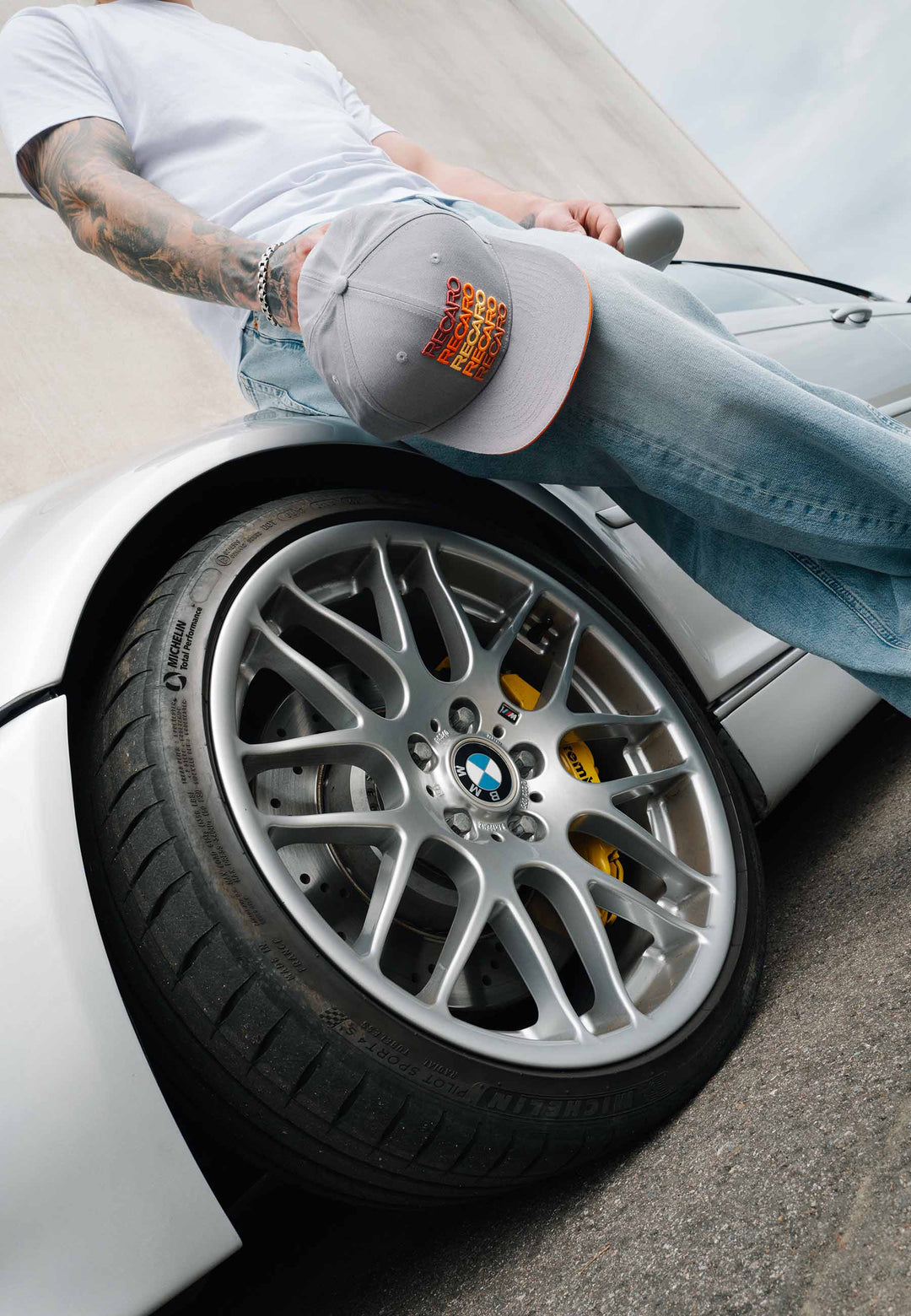 Eine Person in Jeans und einem weißen Hemd steht neben einem silbernen BMW-Sportwagen und hält die RECARO_Merchandising Snapback-Kappe „Spektrum“ in Grau-Orange in der Hand, wobei der Fokus auf den Leichtmetallfelgen und den gelben Bremssätteln im Retro-Stil liegt.