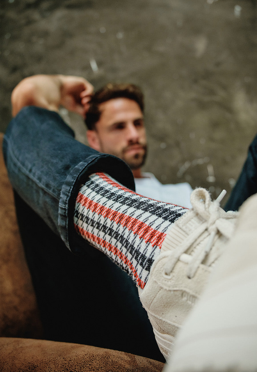 Ein Mann legt entspannt sein Bein hoch und präsentiert einen weißen Sneaker und die Recaro_Merch_Daniel Socken Pepita in einem stilvollen schwarz-weiß-roten Design. Der verschwommene Hintergrund hebt die markante Socke und den Schuh hervor.