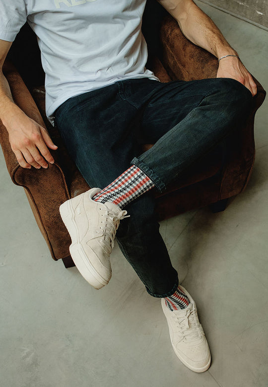 Eine Person sitzt auf einem braunen Stuhl und trägt ein hellblaues T-Shirt, dunkle Jeans, weiße Turnschuhe und Recaro_Merch_Daniel Socken Pepita mit rotem, grauem, schwarzem und weißem Pepita-Muster. Ihre Beine sind auf einem grauen Boden gekreuzt.