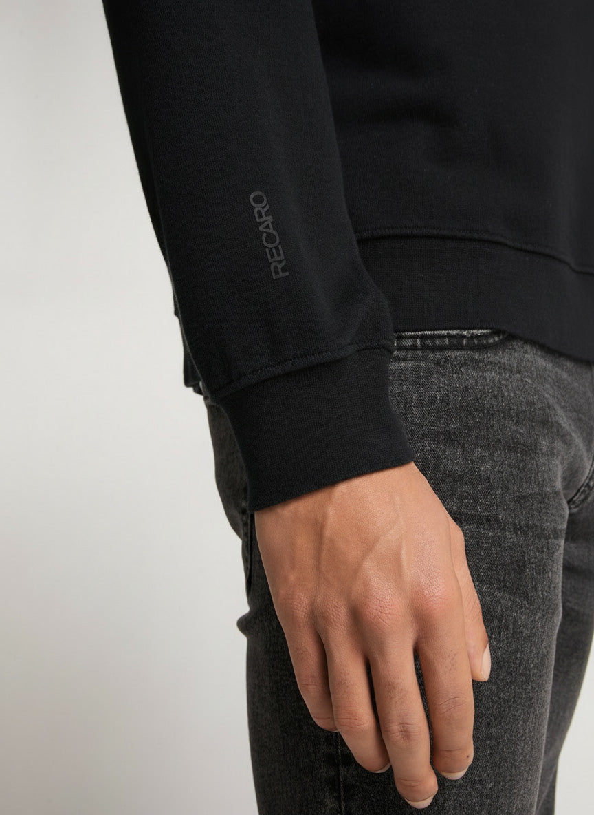 Nahaufnahme der rechten Hand einer Person, die ein schwarzes RECARO  Sweatshirt Originals Rubber Logo mit RECARO-Stickerei auf dem Ärmel trägt, kombiniert mit grauen Relaxed-Fit-Jeans. Der Hintergrund ist schlicht und hell.