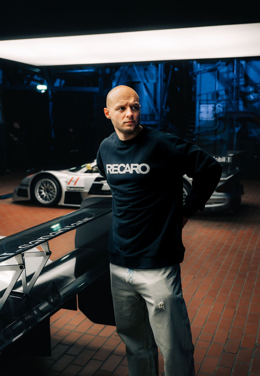 Ein Mann in einem schwarzen RECARO_Merchandising Sweatshirt Originals Rubber Logo und hellen Jeans steht in einem Innenraum auf einem rot gekachelten Boden. Im blau beleuchteten Hintergrund sind Rennwagen und moderne Geräte zu sehen.
