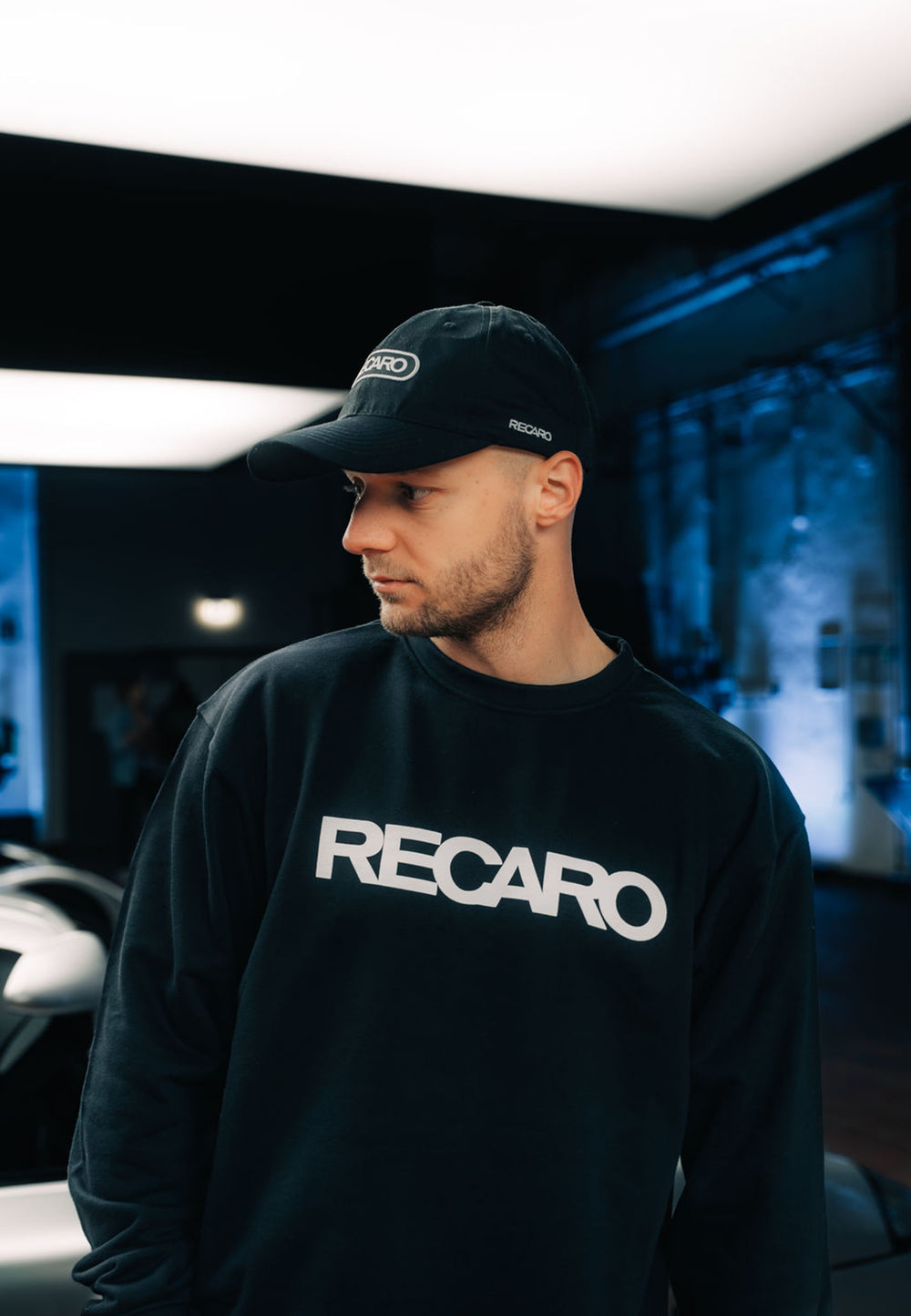 Ein Mann in einem schwarzen RECARO_Merchandising Sweatshirt Originals Rubber Logo steht in einem Innenraum und schaut zur Seite. Blautonige Beleuchtung und verschwommene Industrieelemente unterstreichen den lässigen Stil des Baumwoll-Mischgewebetops.
