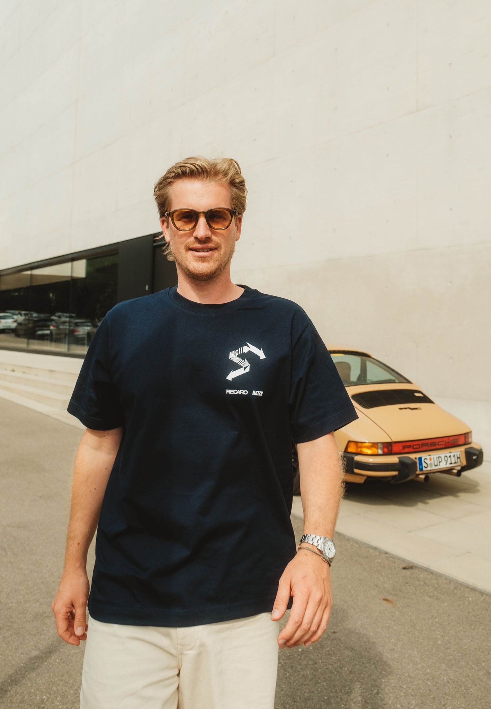 Ein Mann mit Sonnenbrille, hellen Shorts und einem navyfarbenen HEIZR Club X RECARO T-Shirt aus dem RECARO Shop steht im Freien vor einem beigen Oldtimer und einem modernen Betongebäude und zeigt damit authentischen Autokultur-Stil.