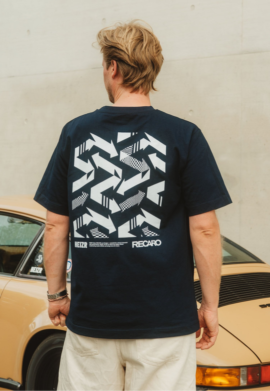 Ein Mann mit blonden Haaren, von hinten gesehen, trägt das T-Shirt HEIZR Club X RECARO von RECARO Shop in Marineblau und hellen Shorts und steht neben einem beigen Oldtimer. Auf dem T-Shirt sind auffällige weiße geometrische Muster und die Schriftzüge "HEIZR" und "RECARO" zu sehen.