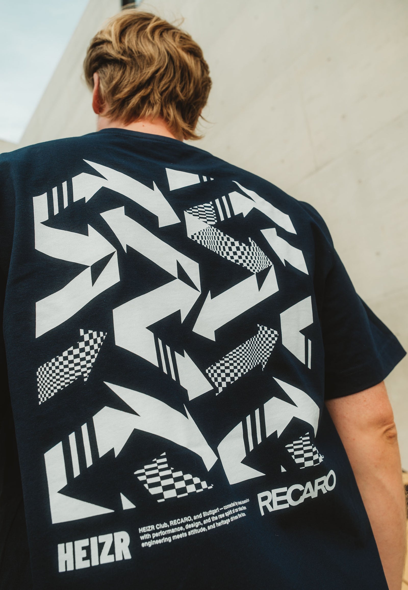 Das T-Shirt HEIZR Club X RECARO aus dem RECARO Shop mit den auffälligen weißen Grafikpfeilen, dem Karomuster und dem HEIZR plus RECARO Schriftzug auf dem Rücken ist ein echtes Highlight für jeden Autokultur-Fan.
