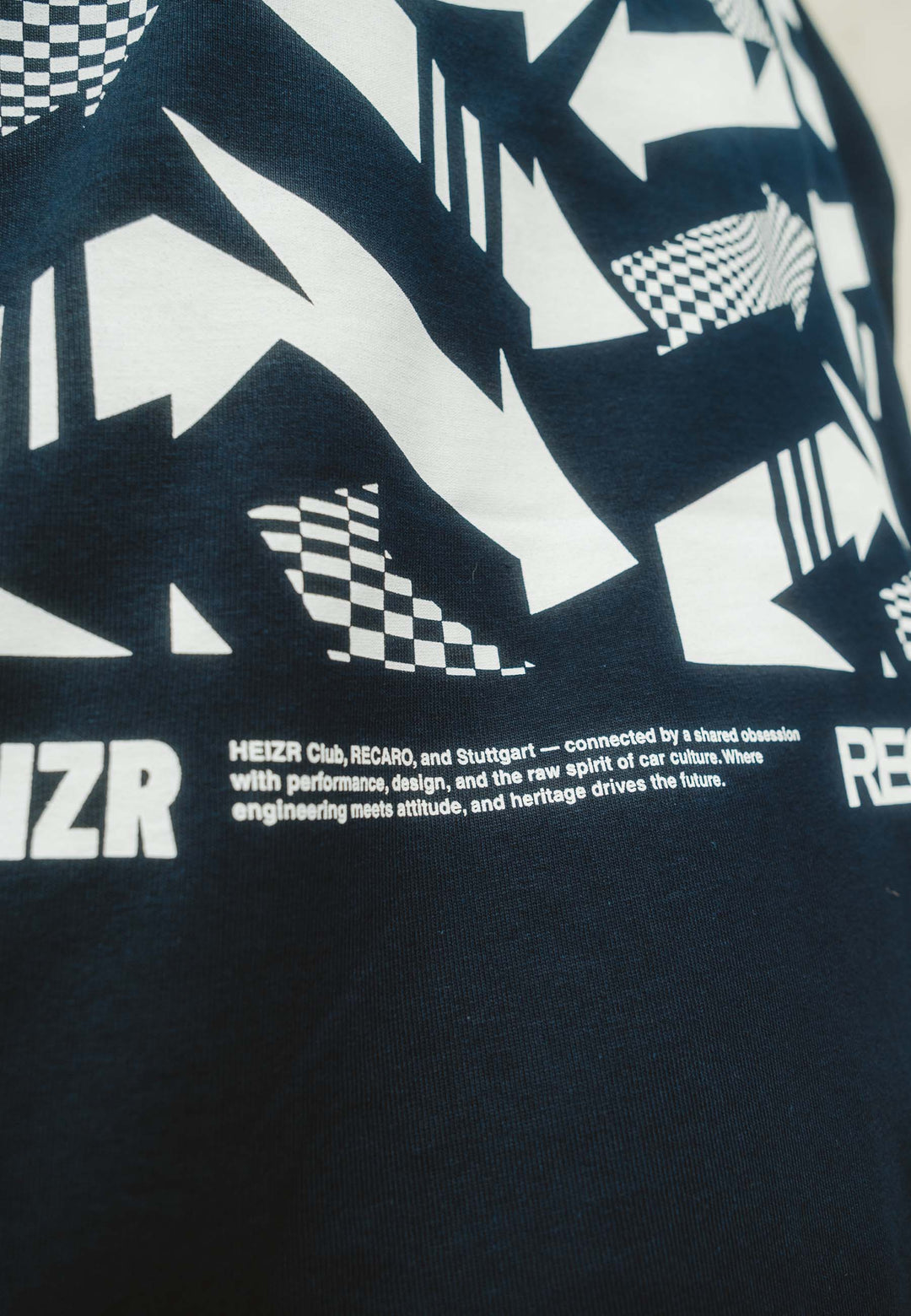 Nahaufnahme des marineblauen T-Shirts HEIZR Club X RECARO aus dem RECARO Shop mit auffälliger weißer Karo-Grafik und Text - HEIZR Club, RECARO, Stuttgart - als Hommage an Autokultur und Performance-Design.