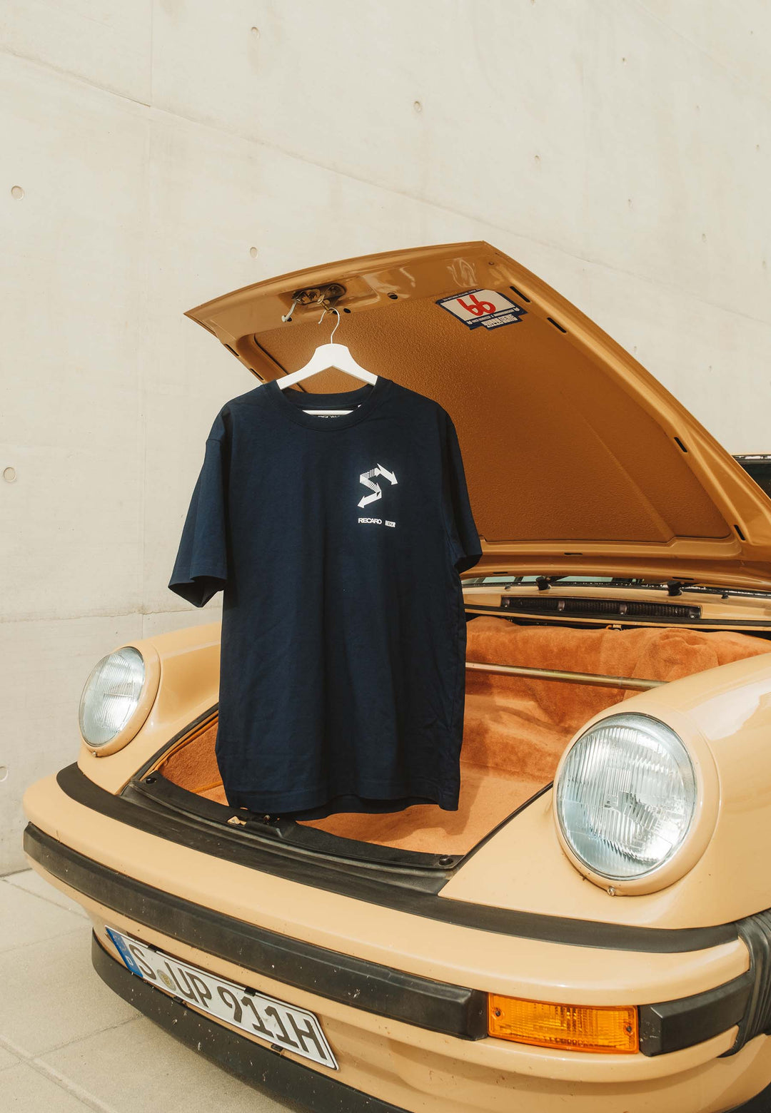 Ein navyfarbenes T-Shirt HEIZR Club X RECARO aus dem RECARO Shop hängt auf einem Bügel am offenen Kofferraum eines beigen Oldtimers mit runden Scheinwerfern, deutschem Nummernschild und hellbraunem, stoffbezogenem Innenraum - ein echter Autokultur-Touch.