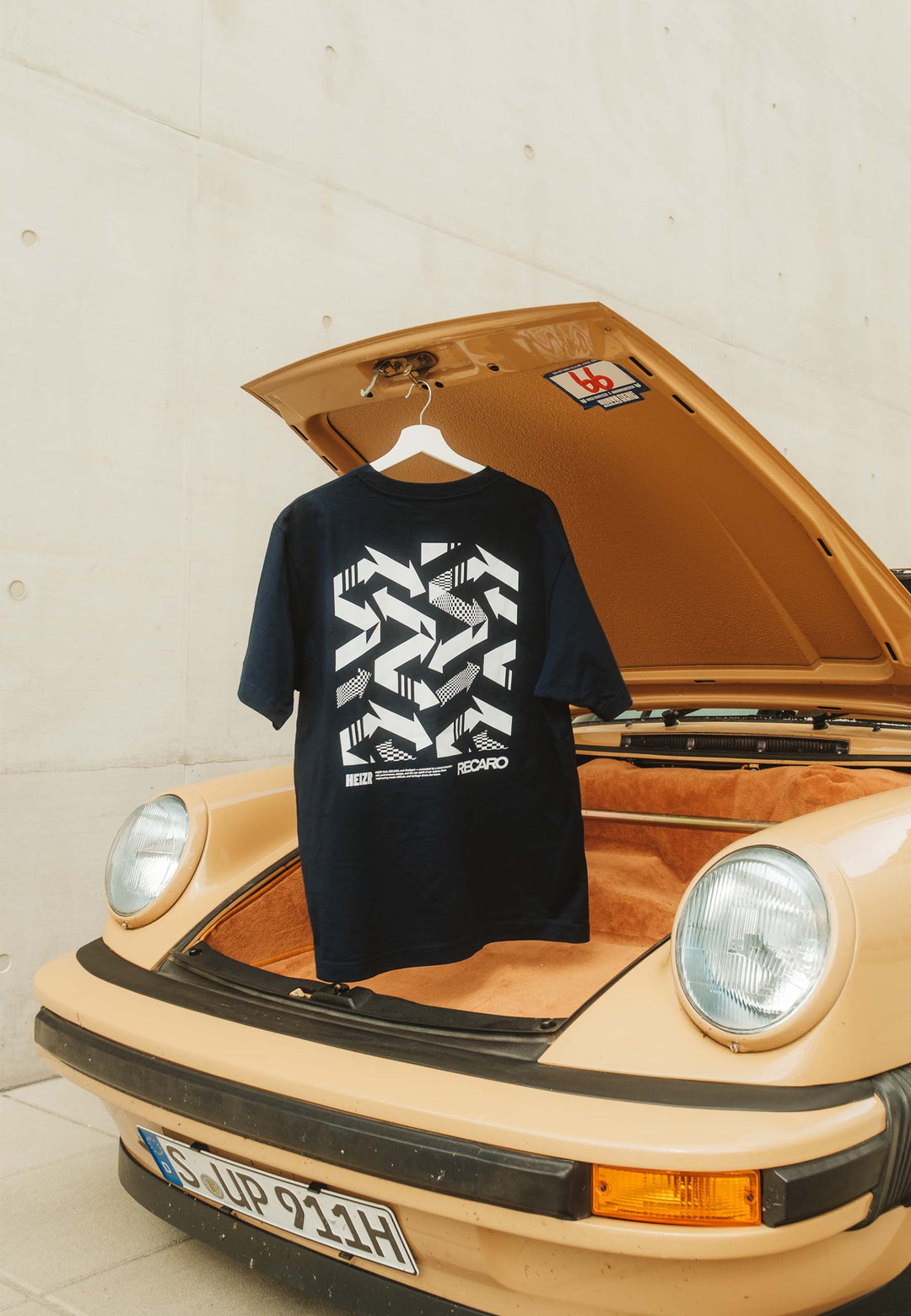 Ein schwarzes T-Shirt HEIZR Club X RECARO aus dem RECARO Shop hängt am offenen Kofferraum eines beigefarbenen Vintage-Porsche, der draußen vor einer minimalistischen Betonwand geparkt ist.