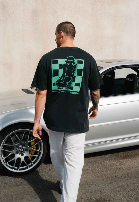 Ein Mann in einem T-Shirt von RECARO Shop mit der Aufschrift „Pole Position“ auf dem Rücken, im Garment-Washed-Look, mit einer grünen Rennsitz-Grafik und einer weißen Hose geht neben einem silbernen Auto mit Leichtmetallfelgen und gelben Bremssätteln her.