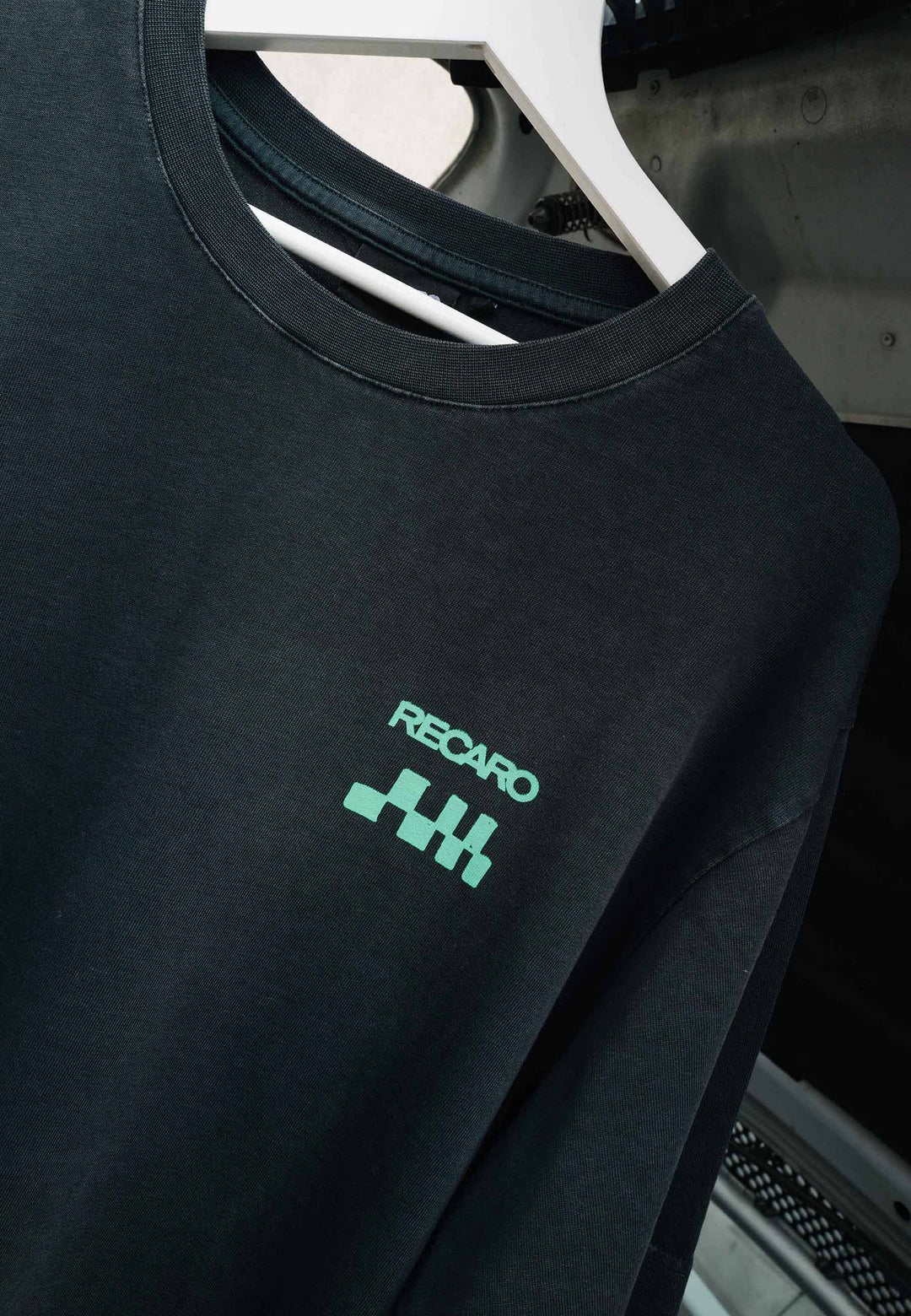 Eine Nahaufnahme eines schwarzen RECARO Shop-T-Shirts mit „Pole Position“-Aufdruck auf der Rückseite, garmentwashed, auf einem weißen Kleiderbügel, mit dem Schriftzug „RECARO“ und einer grünen Rennflagge als Grafik auf der Brust.