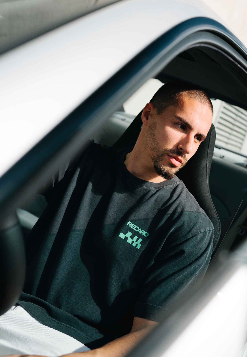 Ein Mann mit kurzen Haaren und einem gepflegten Bart, der ein RECARO Shop-T-Shirt mit der Aufschrift „Pole Position“ auf dem Rücken (garment washed) trägt, sitzt auf dem Fahrersitz eines Autos und blickt nachdenklich aus dem offenen Fenster.
