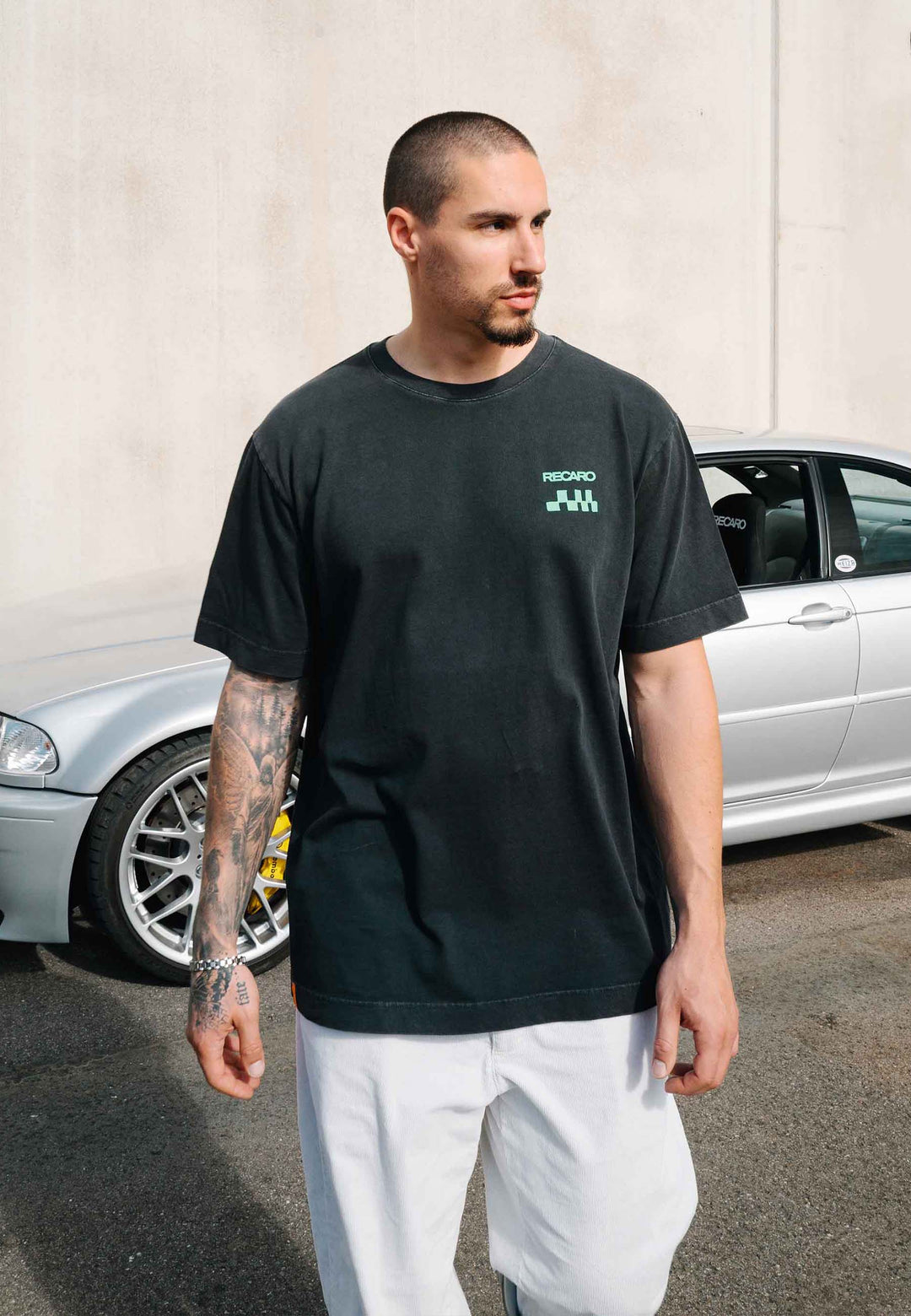 Ein Mann mit rasiertem Kopf und einer Tätowierung am rechten Arm steht draußen neben einem silbernen Auto. Er trägt das schwarze RECARO Shop-T-Shirt „Pole Position Backprint, Garment Washed“ mit grünem Logo sowie eine weiße Hose und blickt zur Seite.