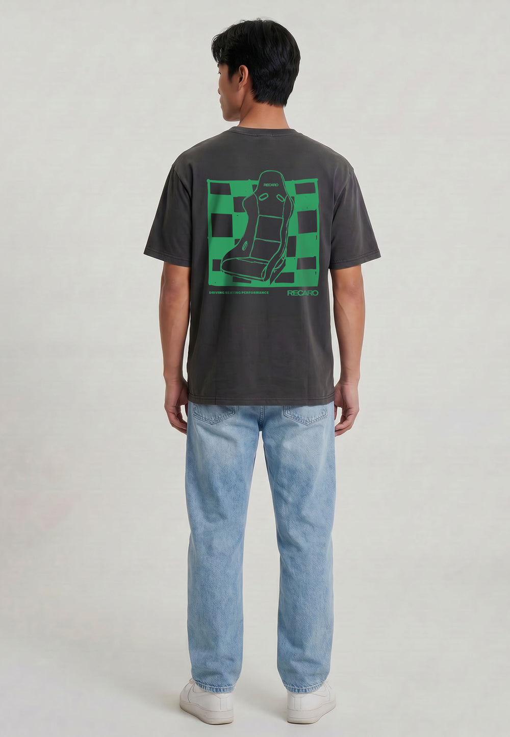 Eine Person blickt weg und trägt das RECARO Shop T-Shirt Pole Position Backprint (Garment Washed) mit einer Autositzgrafik auf grün-kariertem Hintergrund, gepaart mit hellblauen Jeans und weißen Turnschuhen vor einem schlichten Hintergrund.