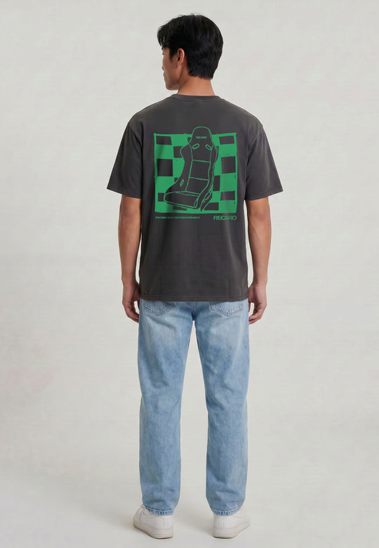 Eine Person blickt weg und trägt das RECARO Shop T-Shirt Pole Position Backprint (Garment Washed) mit einer Autositzgrafik auf grün-kariertem Hintergrund, gepaart mit hellblauen Jeans und weißen Turnschuhen vor einem schlichten Hintergrund.