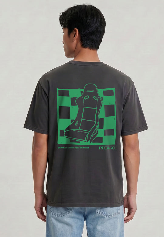 Eine Person blickt weg und trägt das RECARO Shop T-Shirt Pole Position Backprint, Garment Washed, in Dunkelgrau mit grünem Rennsitz und Schachbrettgrafik sowie "RECARO" auf dem Rücken, gepaart mit einer hellblauen Jeans.