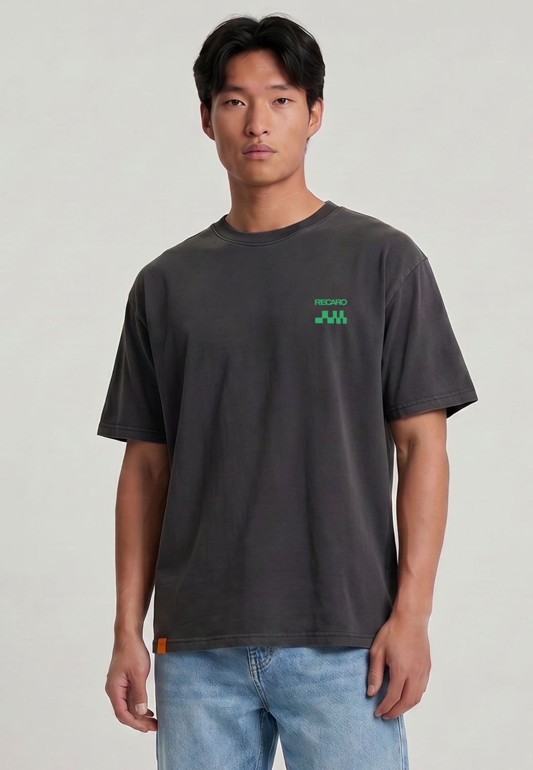 Ein Mann mit glattem schwarzem Haar trägt ein locker sitzendes, dunkelgraues RECARO Shop T-Shirt Pole Position Backprint, Garment Washed, mit kleinem grünem Logo auf der Brust und hellblaue Jeans, vor einem schlichten hellen Hintergrund.