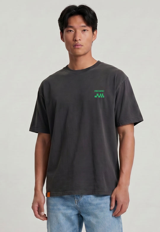Ein Mann mit glattem schwarzem Haar trägt ein locker sitzendes, dunkelgraues RECARO Shop T-Shirt Pole Position Backprint, Garment Washed, mit kleinem grünem Logo auf der Brust und hellblaue Jeans, vor einem schlichten hellen Hintergrund.
