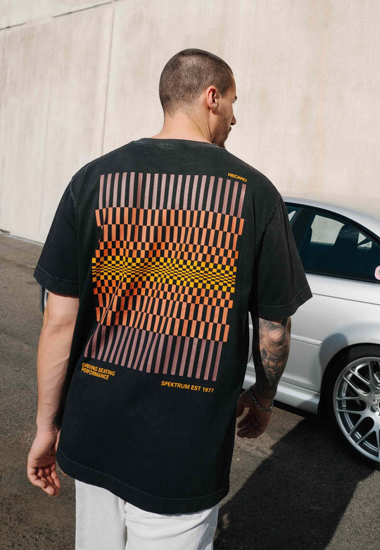 Ein Mann mit kurzen Haaren, von hinten gesehen, trägt das RECARO Shop T-Shirt „Spektrum Backprint“, Garment Washed. Das farbenfrohe geometrische Muster und der Schriftzug fallen besonders ins Auge, während er bei Tageslicht neben einem silbernen Auto vor einer schlichten beigen Wand steht.