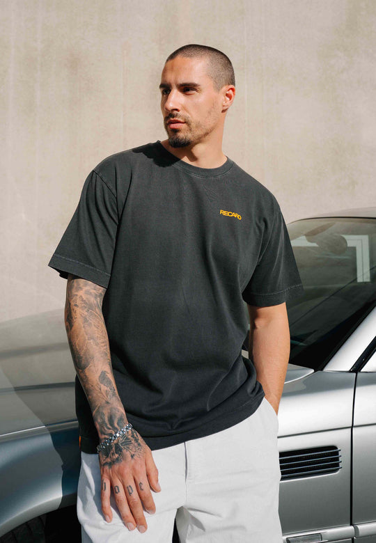 Ein Mann mit kurzen Haaren und Tattoos steht neben einem silbernen Auto. Er trägt das RECARO Shop T-Shirt „Spektrum Backprint, Garment Washed“ – ein übergroßes, schwarzes Unisex-Vintage-T-Shirt mit einem kleinen gelben Logo – sowie weiße Shorts und blickt zur Seite.