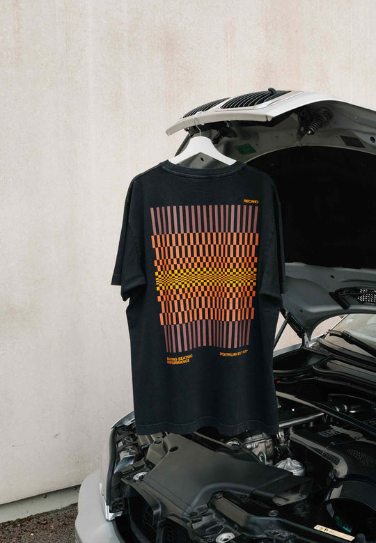 Das RECARO Shop T-Shirt „Spektrum Backprint“, garment-washed, ein übergroßes schwarzes Unisex-Vintage-T-Shirt mit einem orange-roten Aufdruck, liegt über der Motorhaube eines silbernen Autos, das vor einer schlichten hellen Wand geparkt ist.