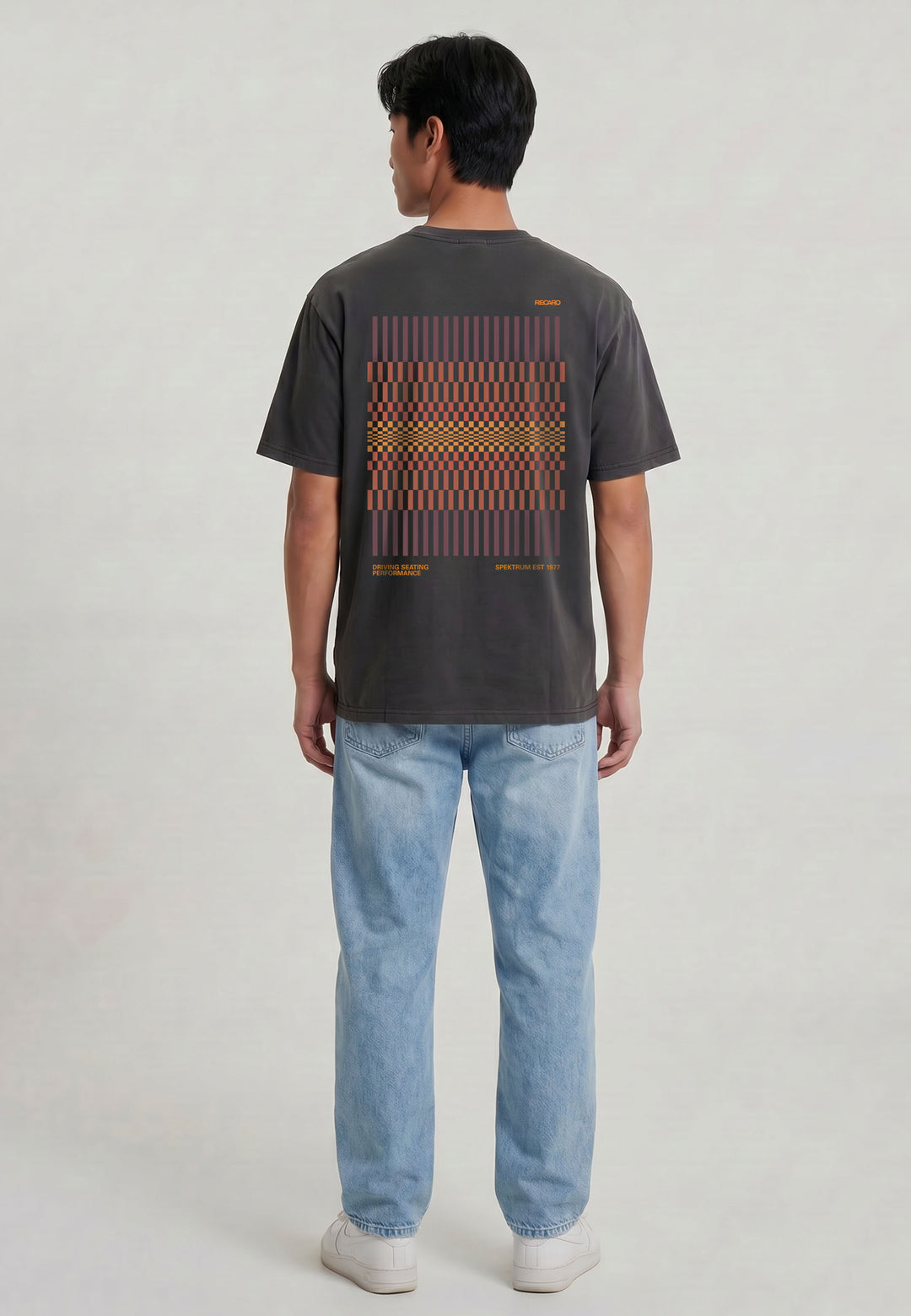 Eine Person steht mit dem RECARO Shop T-Shirt Spektrum Backprint in dunkelgrau (garment washed) mit rotem und orangefarbenem geometrischem Rückendesign, gepaart mit hellblauen Jeans und weißen Turnschuhen vor einem schlichten hellen Hintergrund.