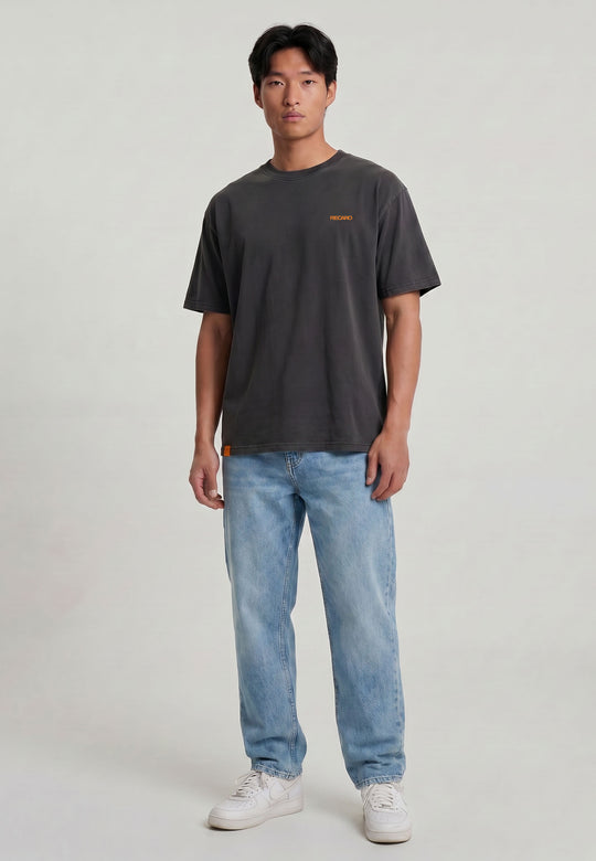 Ein Mann in einem RECARO Shop T-Shirt Spektrum Backprint, Garment Washed (dunkelgrau mit kleinem orangefarbenem Logo), hellblauen Jeans und weißen Turnschuhen posiert vor einem schlichten hellen Hintergrund.