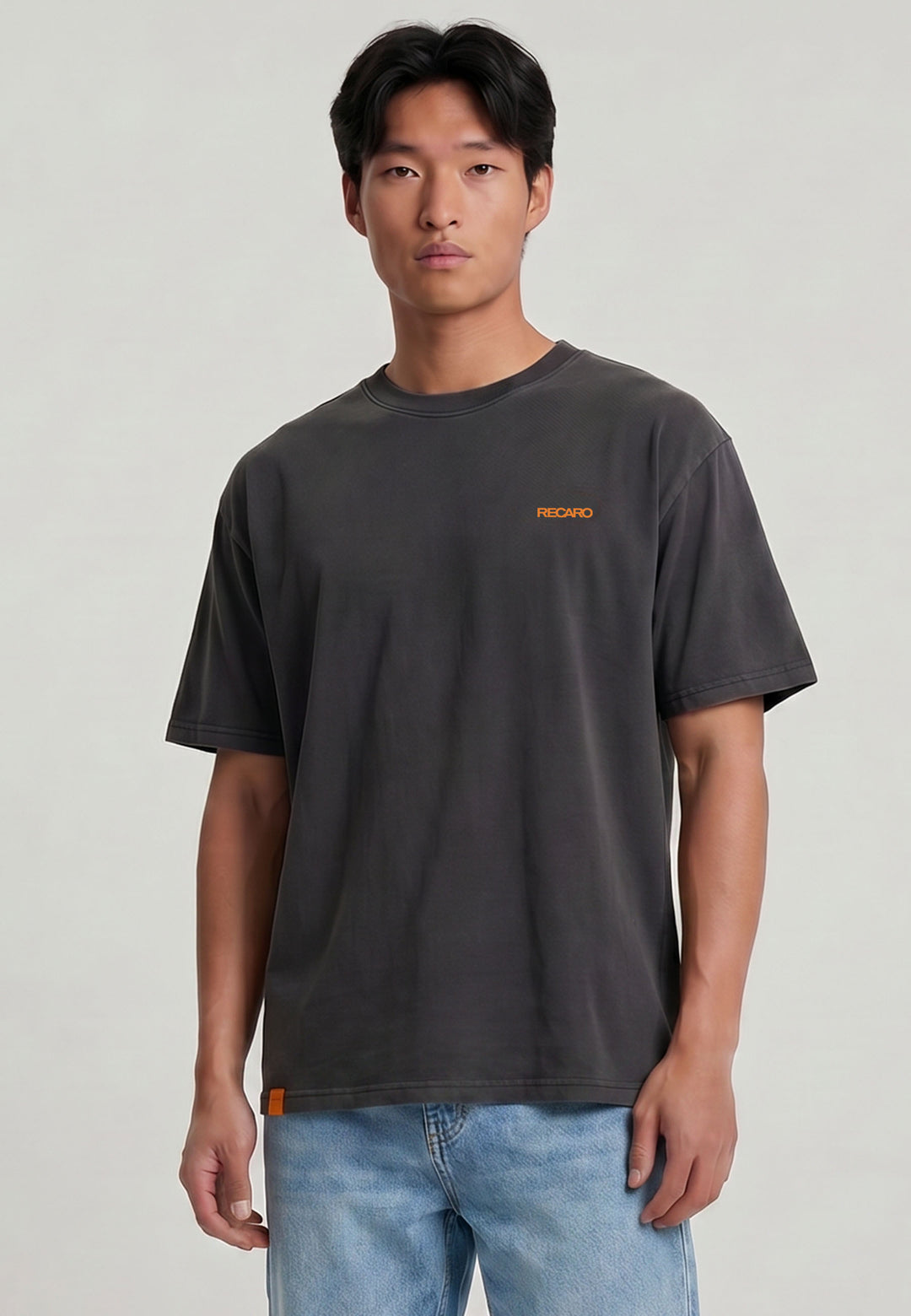 Ein junger Mann mit kurzen schwarzen Haaren trägt das RECARO Shop T-Shirt Spektrum Backprint, Garment Washed - ein lockeres dunkelgraues T-Shirt mit RECAND in kleinen orangefarbenen Buchstaben auf der Brust - zusammen mit einer hellblauen Jeans vor einem schlichten hellen Hintergrund.