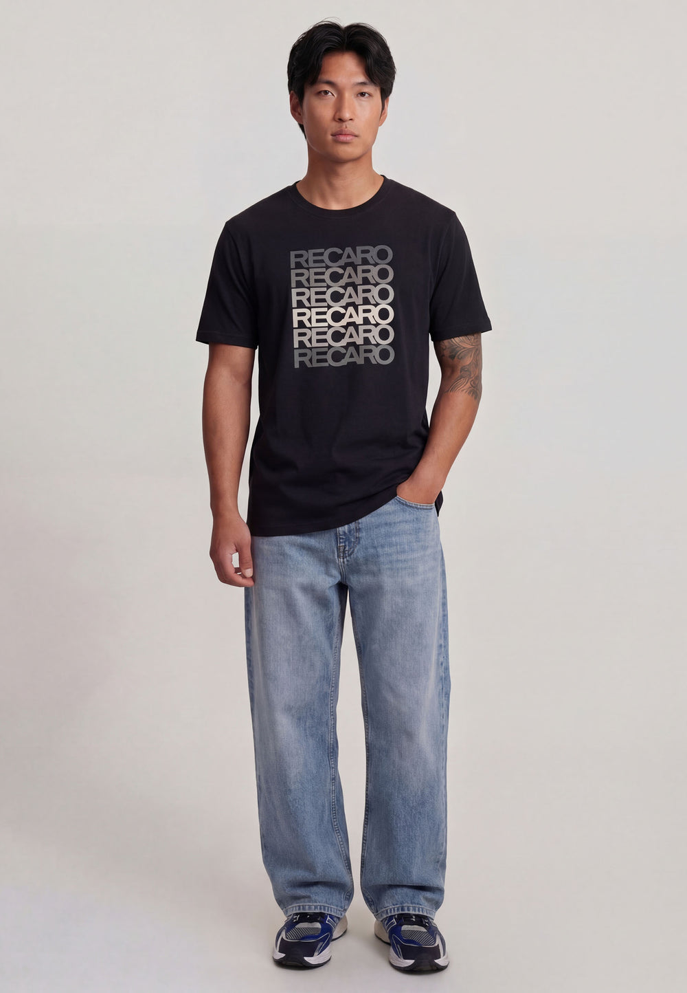 Ein Mann mit kurzen schwarzen Haaren trägt ein Recaro_Merch_Daniel T-Shirt Spektrum grau, hellblaue Jeans und blaue Turnschuhe und steht vor einem schlichten hellen Hintergrund.