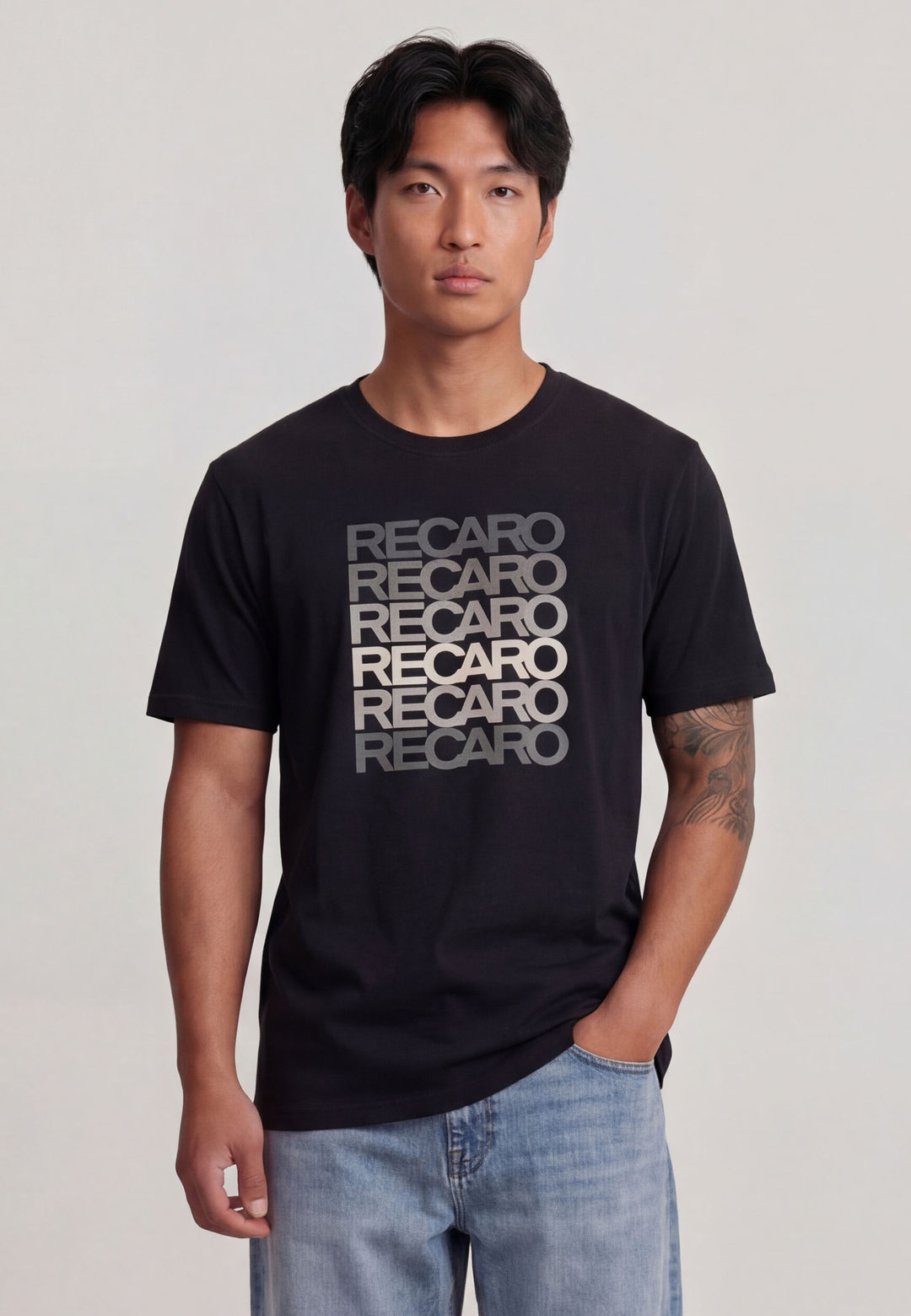 Ein Mann mit schwarzen Haaren trägt das "T-Shirt Spektrum grey" von Recaro_Merch_Daniel mit wiederholtem grauem Text auf weicher Baumwolle, gepaart mit einer blauen Jeans vor einem schlichten hellen Hintergrund.