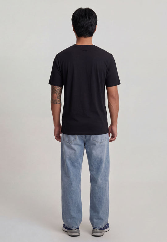 Eine Person mit kurzen dunklen Haaren steht mit dem Gesicht nach hinten, trägt das schwarze T-Shirt Spektrum von Recaro_Merch_Daniel, hellblaue Jeans und Turnschuhe. Ihr tätowierter rechter Arm ist vor einem schlichten hellen Hintergrund im Retro-Look zu sehen.