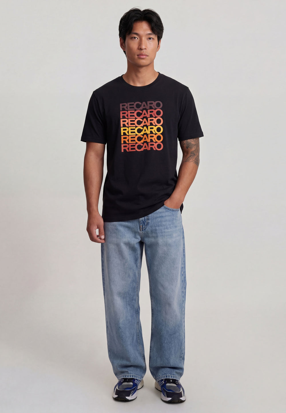 Ein Mann blickt nach vorne in einem schwarzen Recaro_Merch_Daniel T-Shirt Spektrum mit "RECARO" in Orange und Rot, gepaart mit hellblauen Jeans und dunklen Turnschuhen für einen coolen Retro-Look vor einem schlichten hellen Hintergrund.