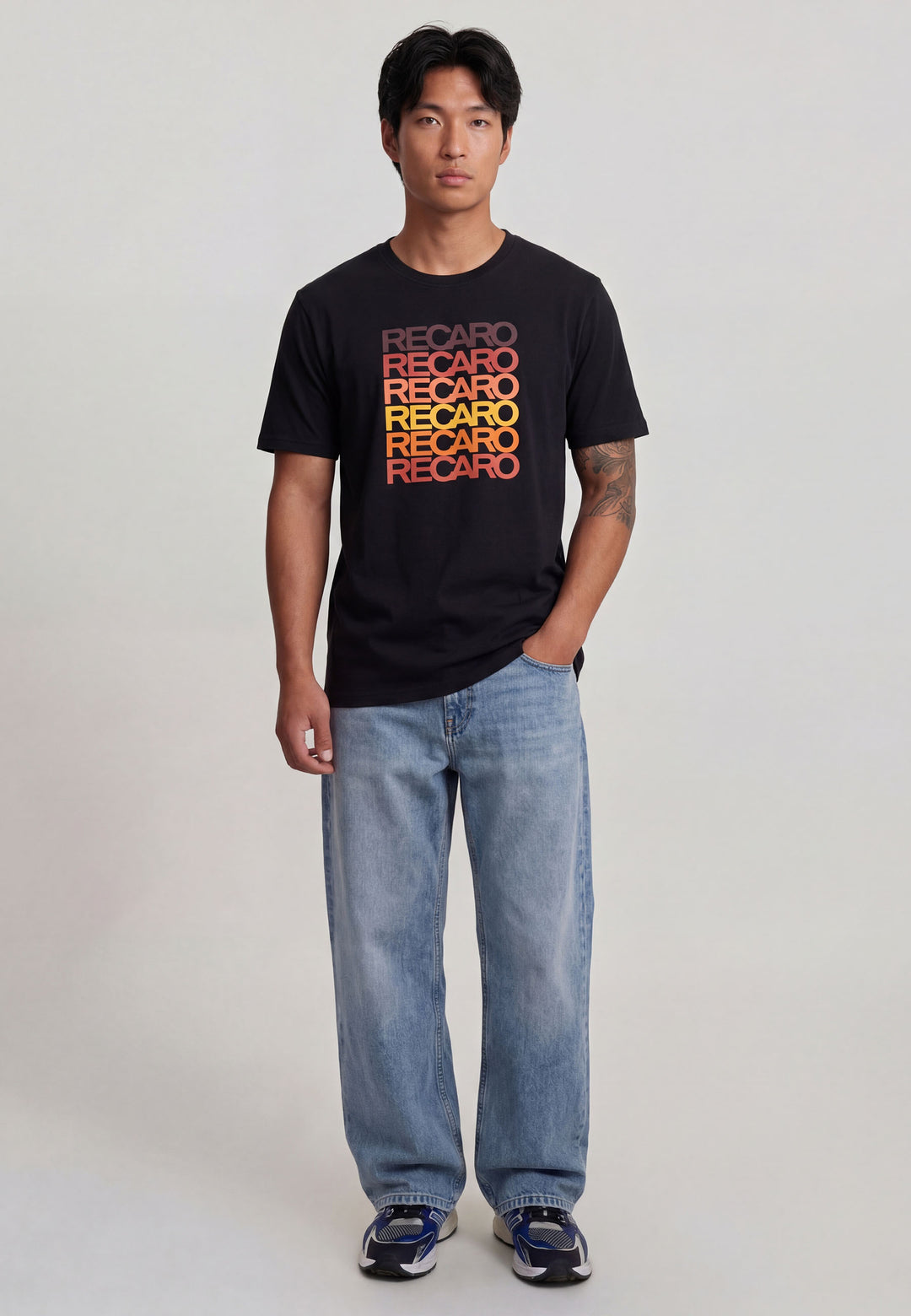Ein Mann blickt nach vorne in einem schwarzen Recaro_Merch_Daniel T-Shirt Spektrum mit "RECARO" in Orange und Rot, gepaart mit hellblauen Jeans und dunklen Turnschuhen für einen coolen Retro-Look vor einem schlichten hellen Hintergrund.
