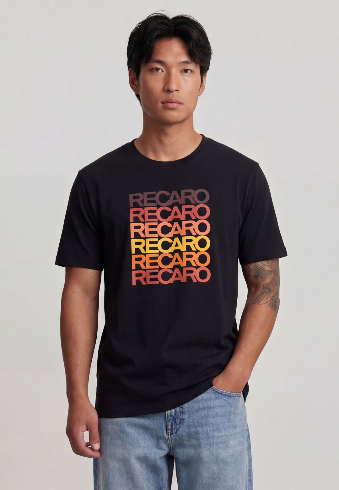 Ein Mann mit kurzen schwarzen Haaren trägt das Recaro_Merch_Daniel T-Shirt Spektrum schwarz, aus Baumwolle, mit einem sich wiederholenden RECARO-Aufdruck im Retro-Look in einem Farbverlauf von rot nach gelb. Er steht mit einer Hand in seiner Jeanstasche vor einem unifarbenen Hintergrund.