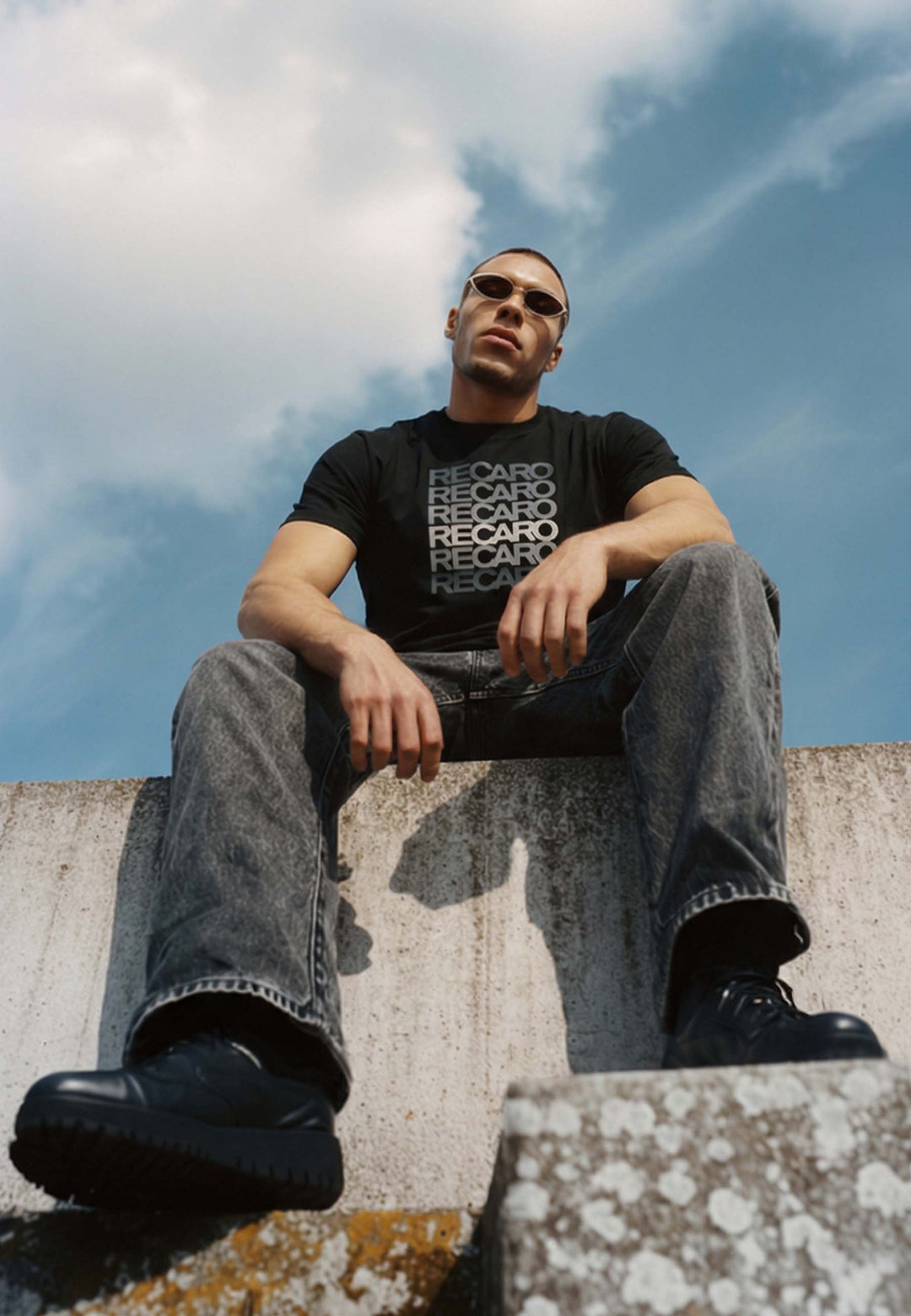 Ein Mann im Retro-Look, mit Sonnenbrille und einem Recaro_Merch_Daniel T-Shirt Spektrum grau aus 100% Baumwolle, grauen Jeans und schwarzen Schuhen sitzt auf einem Betonvorsprung und blickt auf einen blauen Himmel mit Wolken.