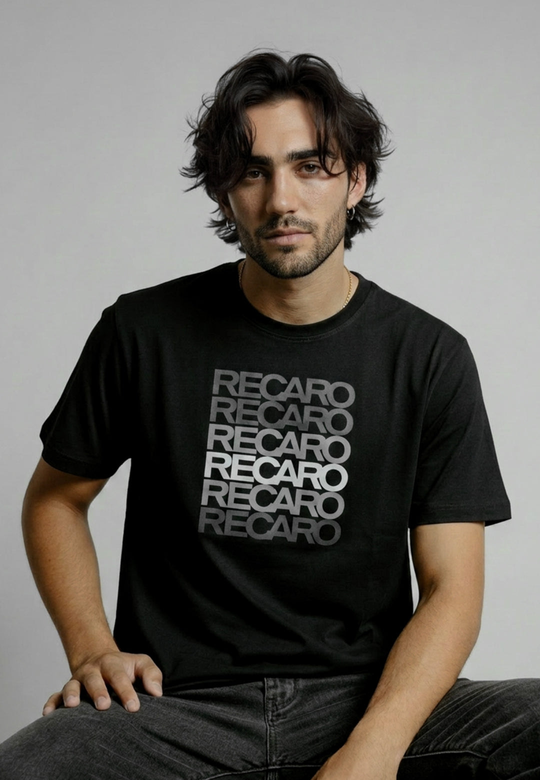 Ein Mann mit gewelltem dunklem Haar und Gesichtsbehaarung sitzt vor einem schlichten Hintergrund und trägt das Recaro_Merch_Daniel T-Shirt Spektrum grau. Das Shirt ist mit einem fetten weißen Schriftzug versehen, und er wirkt in einer lässigen Pose entspannt.