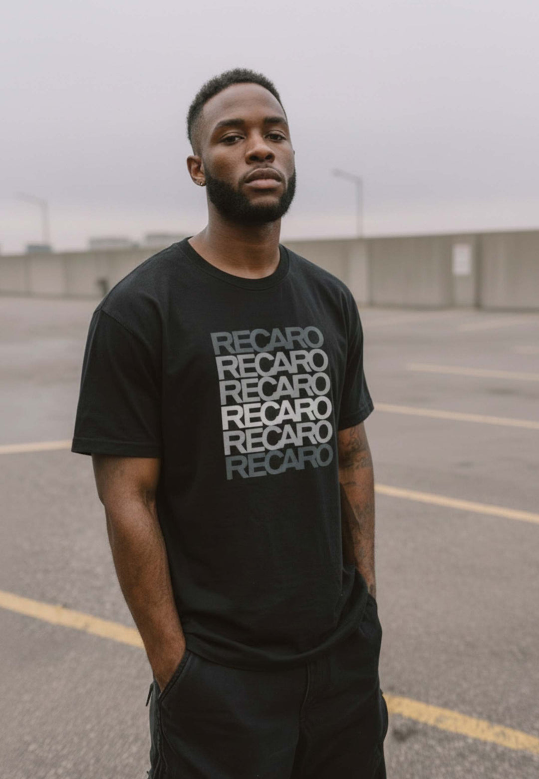 Ein Mann mit kurzen Haaren und einem Bart steht auf einem leeren Parkplatz, die Hände in den Taschen, und trägt das Recaro_Merch_Daniel T-Shirt Spektrum grau. Der bedeckte Himmel unterstreicht die klassische Ausstrahlung dieses Baumwoll-T-Shirts im Retro-Look mit grauem Schriftzug.