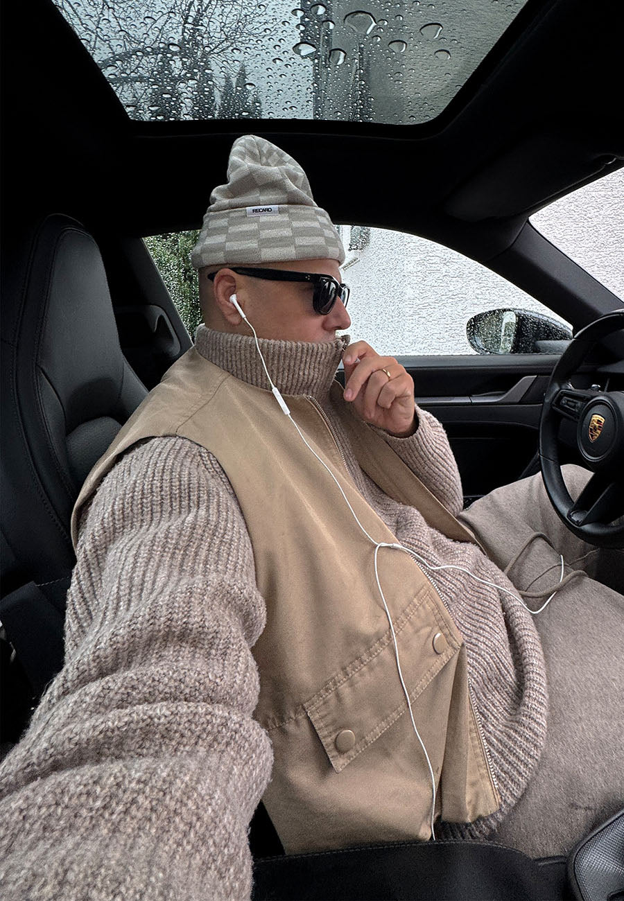 Eine Person, die eine beige RECARO_Merchandising Beanie Checkered, eine Sonnenbrille, eine beige Weste und einen dicken Strickpullover trägt, sitzt mit Kopfhörern auf dem Fahrersitz des Autos. Regen trübt das Schiebedach, draußen sind Bäume zu sehen.