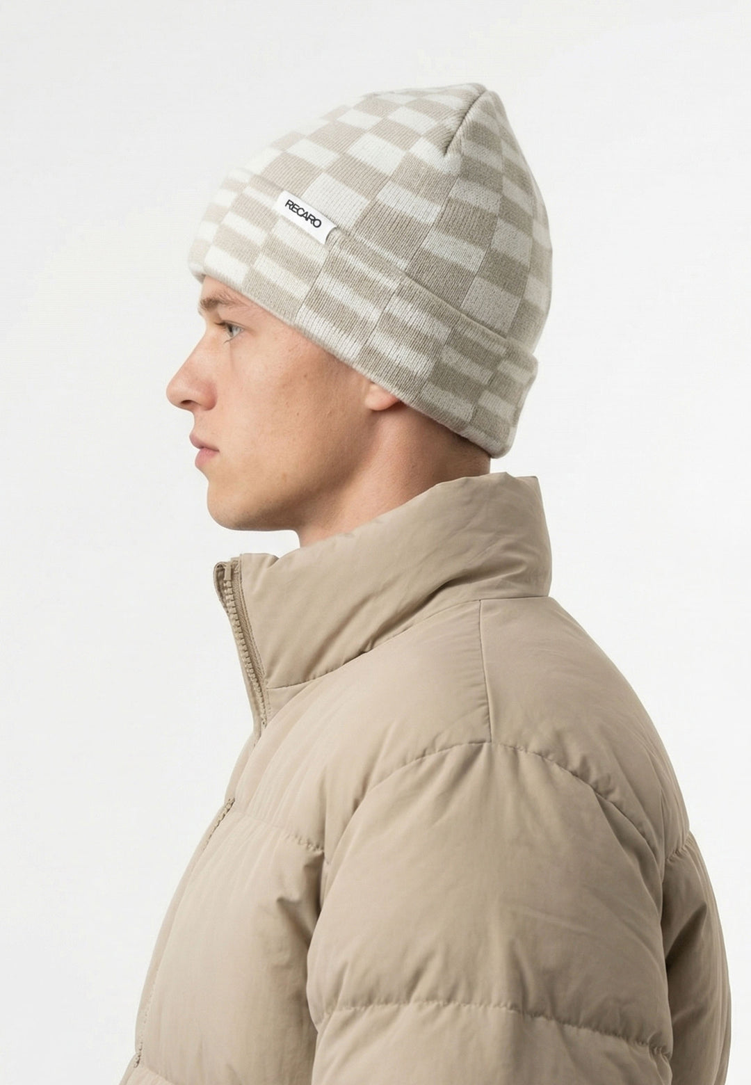 Eine Person in beiger Pufferjacke und hellgrau-weißer RECARO_Merchandising Beanie Checkered, nach links blickend vor einem schlichten, hellen Hintergrund.
