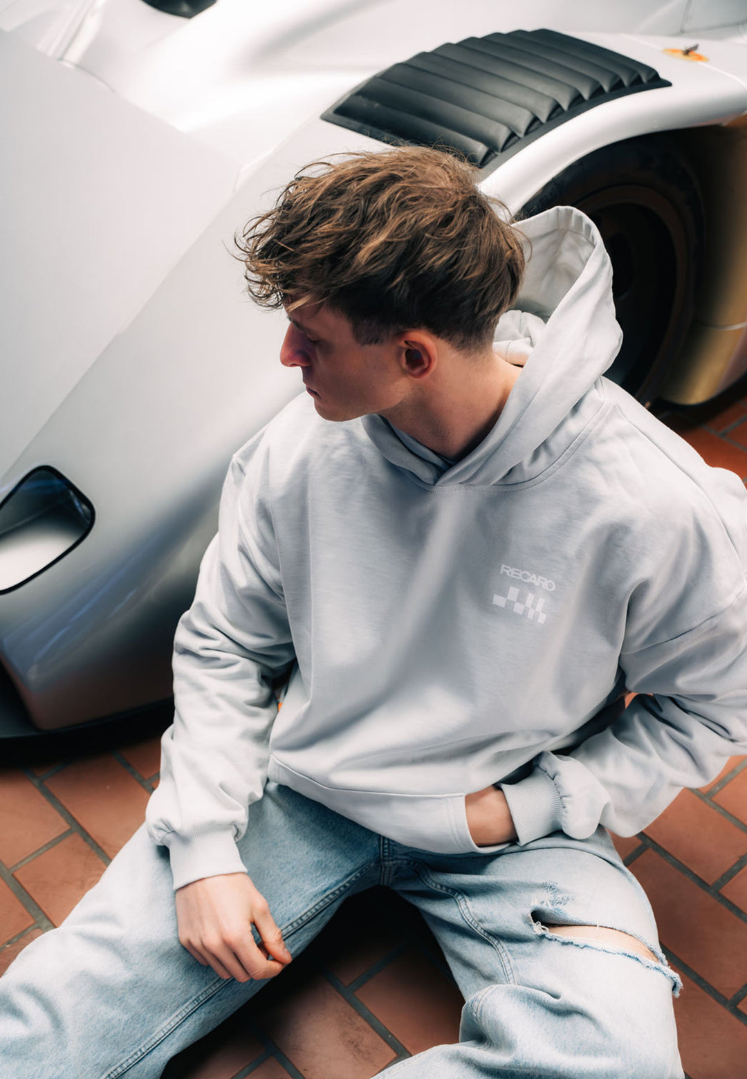 Ein junger Mann in einem übergroßen hellgrauen Hoodie RECARO Heavy Cotton by RECARO Shop und zerrissenen Jeans sitzt auf einem Fliesenboden neben einem schnittigen silbernen Sportwagen und schaut entspannt nach links.