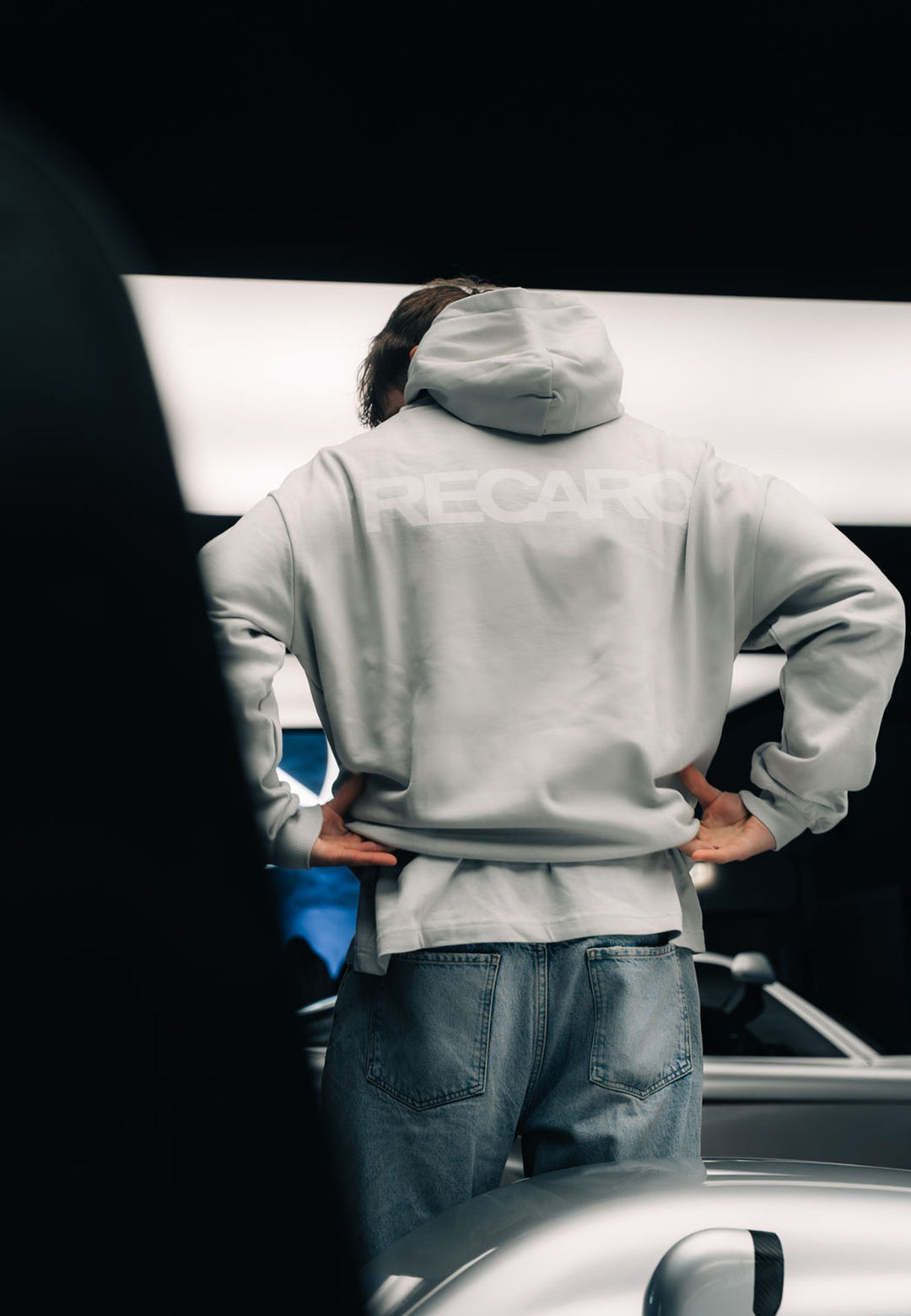 Eine Person in einem übergroßen hellgrauen Hoodie RECARO Heavy Cotton aus dem RECARO Shop steht mit den Händen in den Hüften und dem Blick abgewandt neben einem silbernen Auto in einem Innenraum bei heller Beleuchtung.