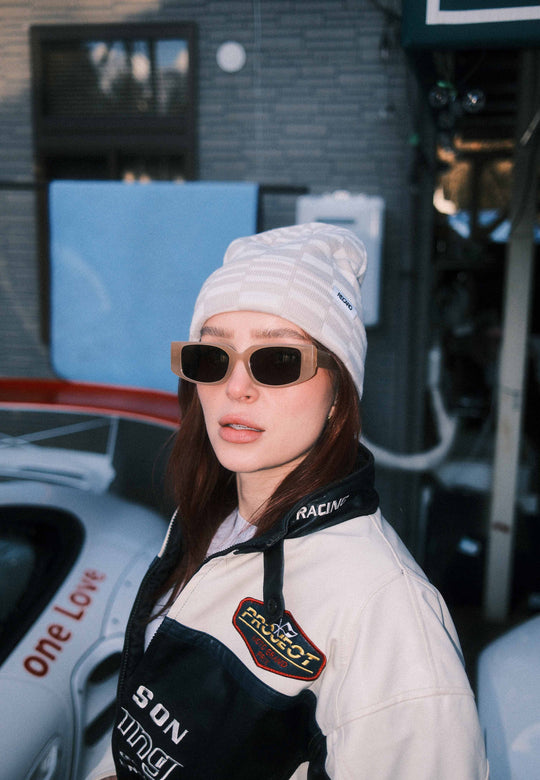 Eine Frau mit RECARO_Merchandising Beanie Checkered, Sonnenbrille und Rennjacke steht neben einem weißen Sportwagen mit der Aufschrift "One Love"; im Hintergrund sind Gebäude und Geräte zu sehen.
